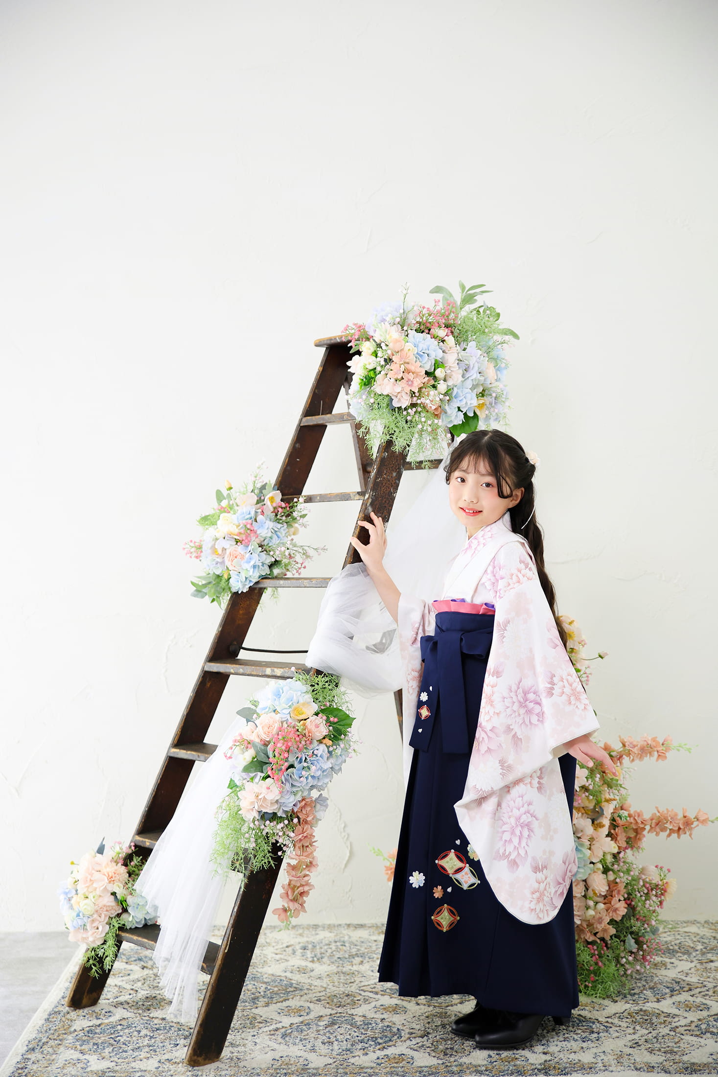 白地に淡いピンクの花柄が描かれた着物と、紺色の袴を合わせた女性の記念写真。髪には白い髪飾りが添えられ、両手には小さな花を持っている。背景や手前には淡い紫色の藤の花が美しく垂れ下がり、柔らかく明るい雰囲気を演出している。卒業や記念撮影にふさわしい、可憐で春らしい一枚。