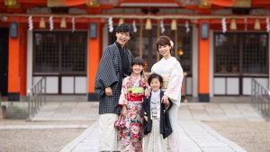 七五三の由来と意味、お祝いの時期や方法は? 朱色の神社を背景に、着物や羽織袴姿で家族仲良く笑顔並んで立つ七五三の記念撮影写真