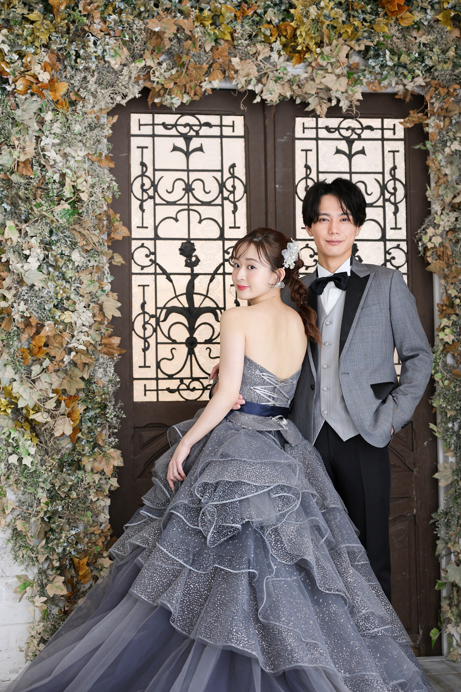 カラードレスとタキシードでフォトウェディングの撮影をする新郎新婦様。ナチュラルな蔦に囲まれた、鉄格子デザインのアンティーク扉を背景に撮影している写真。撮影店舗：しんかな大阪南店 住所：大阪府堺市北区長曽根町3069-7