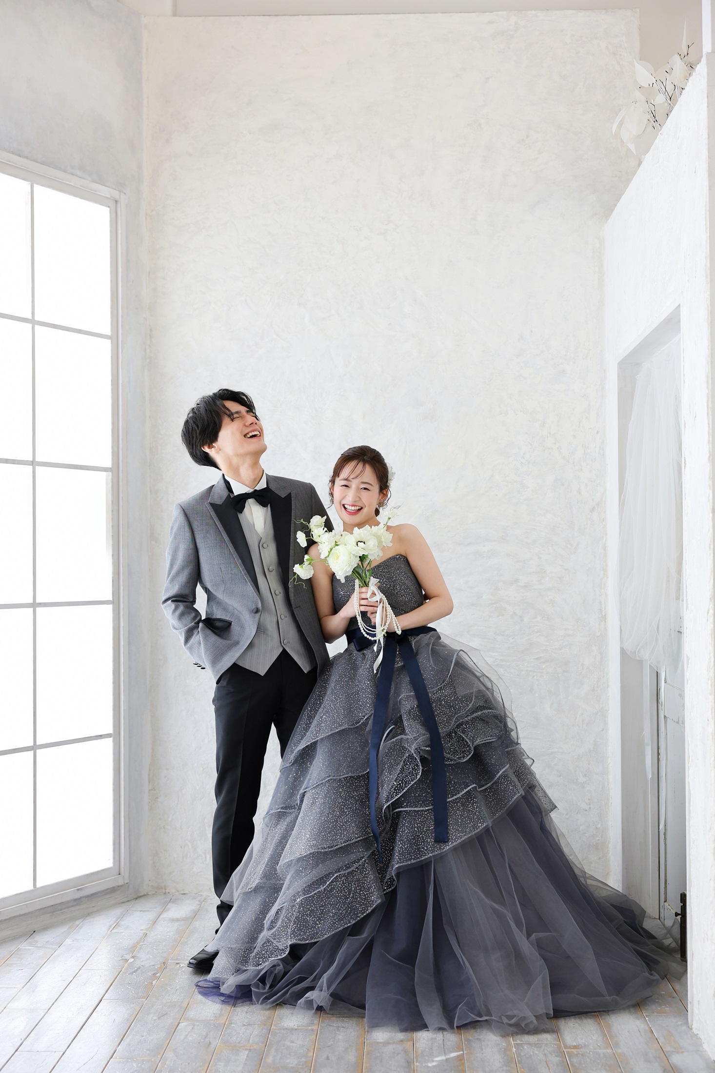 カラードレスとタキシードでフォトウェディングの撮影をする新郎新婦様。大きな窓から降り注ぐ光に包まれる中、グレー色の淡いカラードレスが映える新婦様と新郎様を撮影している写真。撮影店舗：しんかな大阪南店 住所：大阪府堺市北区長曽根町3069-7