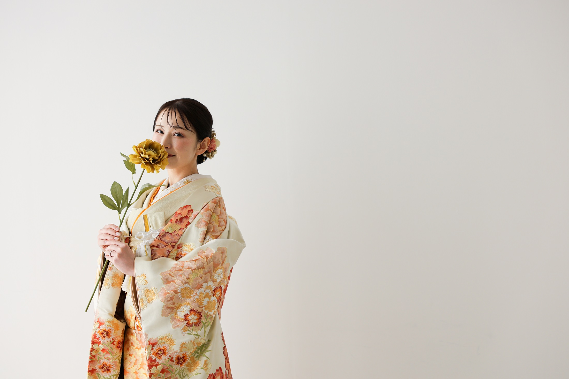 色打掛と袴でフォトウェディングの撮影をする新郎新婦様。真っ白の壁を背景に、一輪のお花を持ち、オレンジ色の色打掛が映える新婦様を撮影している写真。撮影店舗：しんかな大阪南店 住所：大阪府堺市北区長曽根町3069-7
