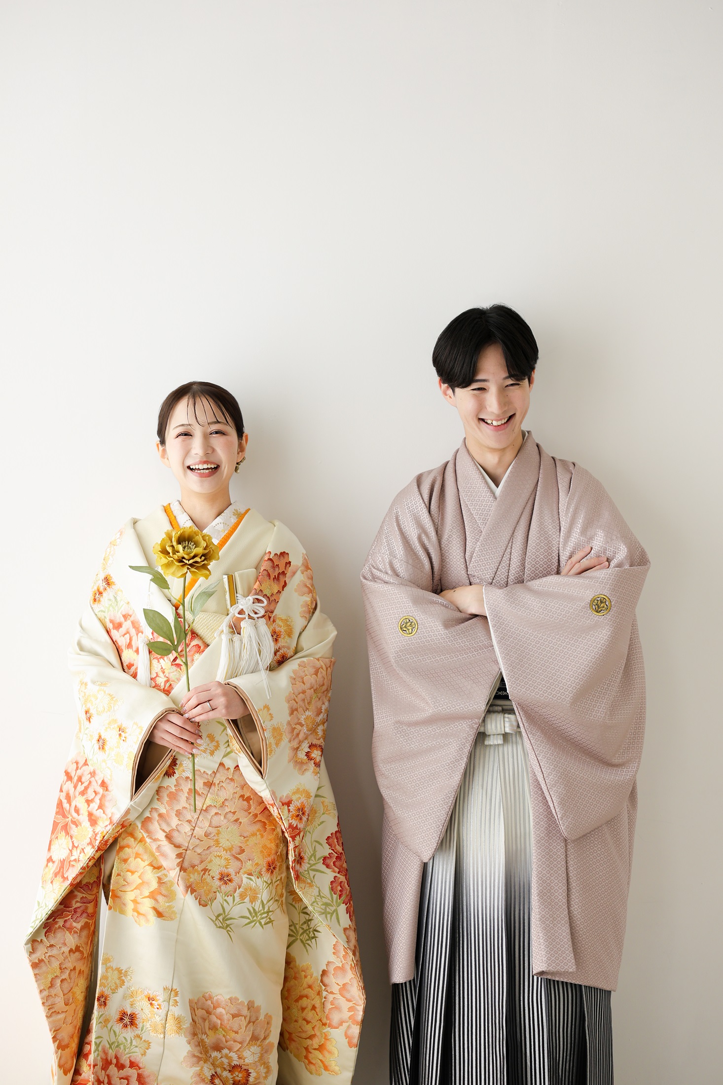 色打掛と袴でフォトウェディングの撮影をする新郎新婦様。真っ白の壁を背景に、オレンジ色が映える色打掛の新婦様が一輪のお花を持ち、新郎様と並んで撮影している写真。オレンジ大きな窓から降り注ぐ光に包まれ撮影をする新郎新婦様。撮影店舗：しんかな大阪南店 住所：大阪府堺市北区長曽根町3069-7