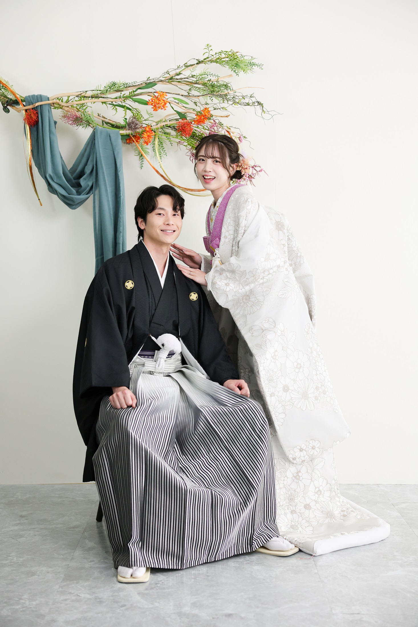 色打掛と袴でフォトウェディングの撮影をする新郎新婦様。真っ白の壁に、オレンジの花や葉、緑色のカーテンを飾りつけたスタイリッシュなハンギングブランチを背景に撮影している写真。撮影店舗:芝ParksideSTUDIO店 住所:東京都港区芝2-1-28 芝アネックスビル1階