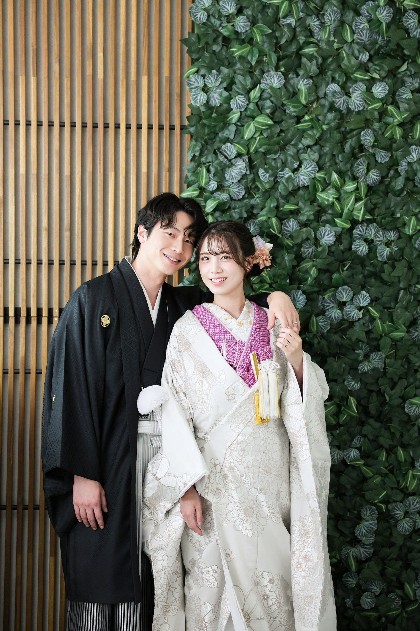 色打掛と袴でフォトウェディングの撮影をする新郎新婦様。緑の葉や木製の壁を背景に、ピンク色の襟元が映える新婦様と新郎様を撮影している写真。撮影店舗:芝ParksideSTUDIO店 住所:東京都港区芝2-1-28 芝アネックスビル1階