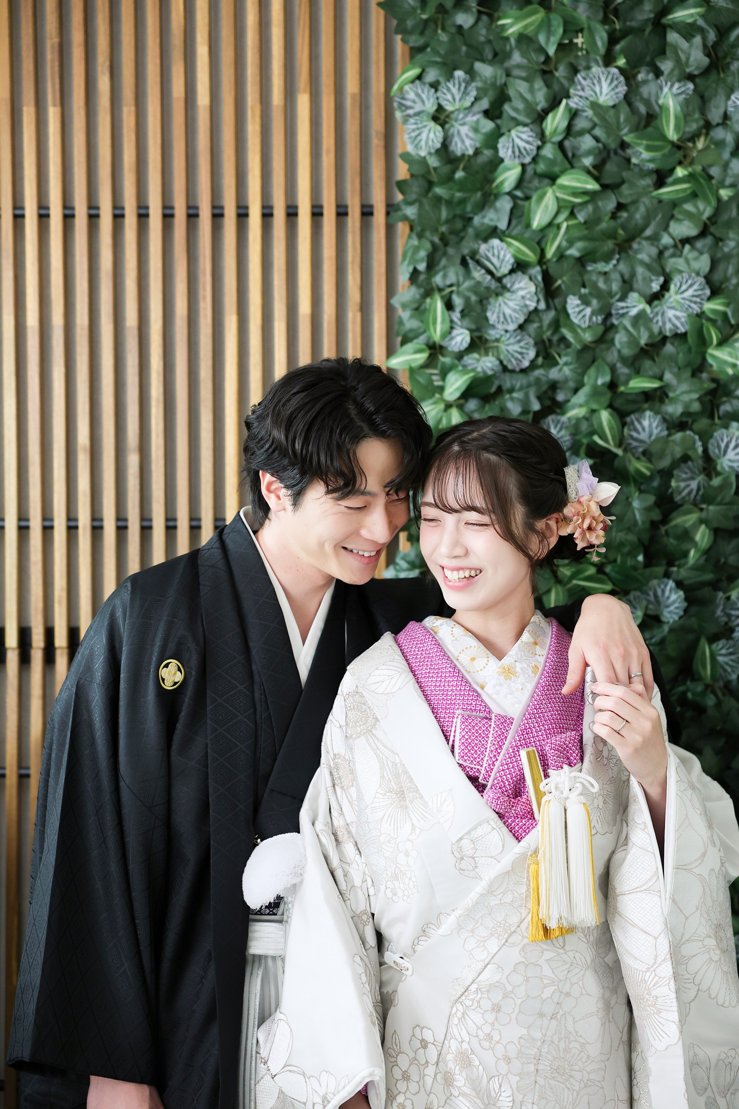 色打掛と袴でフォトウェディングの撮影をする新郎新婦様。緑の葉や木製の壁を背景に、ピンク色の襟元が映える新婦様と新郎様を撮影している写真。撮影店舗:芝ParksideSTUDIO店 住所:東京都港区芝2-1-28 芝アネックスビル1階