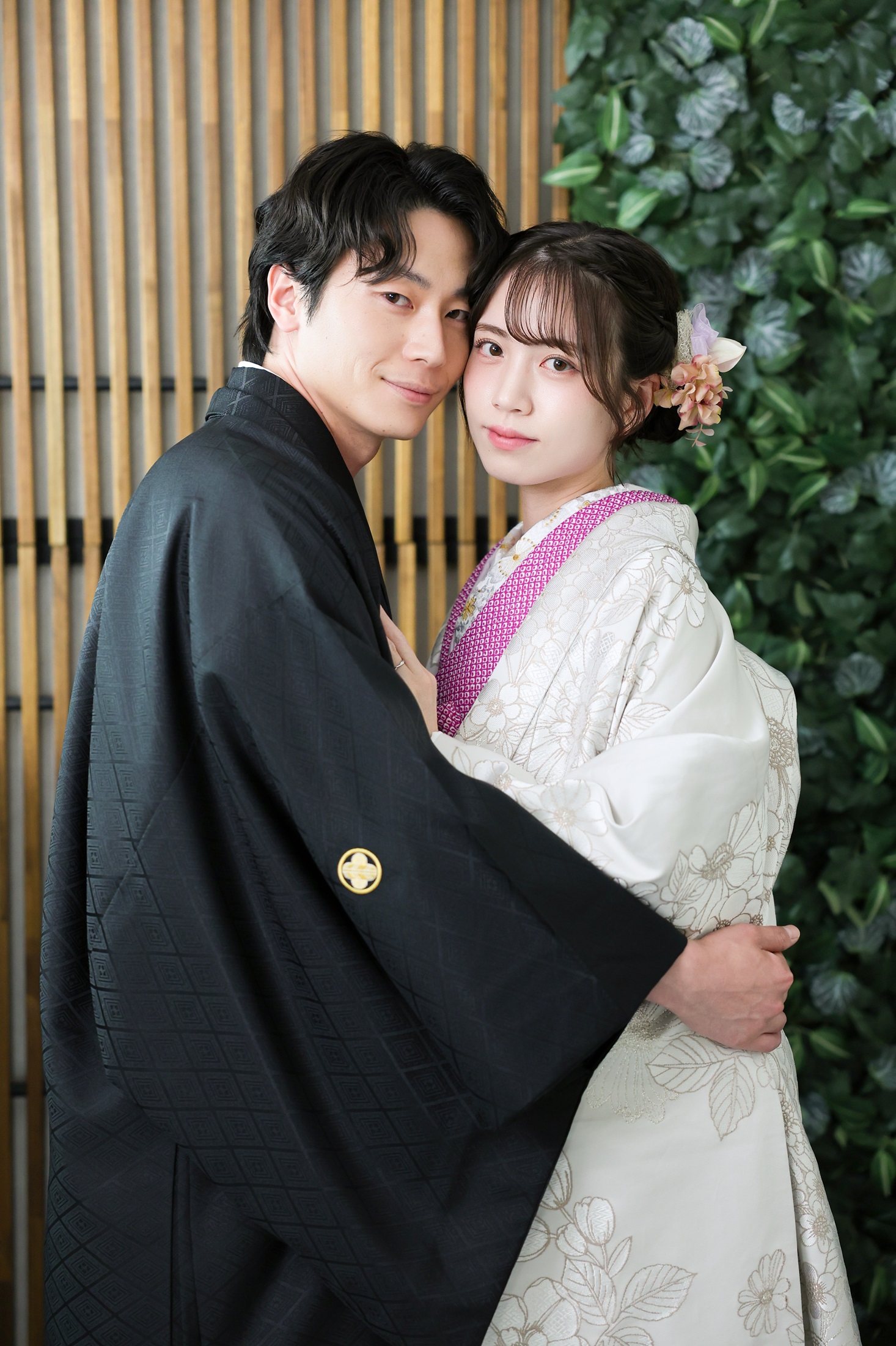 色打掛と袴でフォトウェディングの撮影をする新郎新婦様。緑の葉や木製の壁を背景に、ピンク色の襟元が映える新婦様と新郎様を撮影している写真。撮影店舗:芝ParksideSTUDIO店 住所:東京都港区芝2-1-28 芝アネックスビル1階