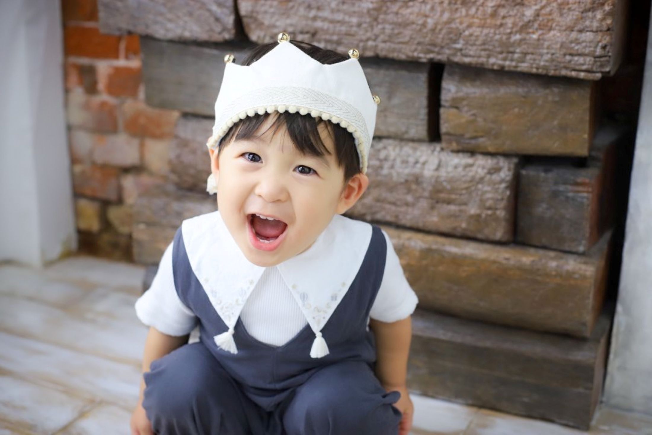 七五三3歳男の子の記念写真。レンガの壁や木材を背景に、青いオーバーオールに白い生地の王冠をかぶり、王子様のような白い襟で、しゃがんでとびきりの笑顔を見せる写真。