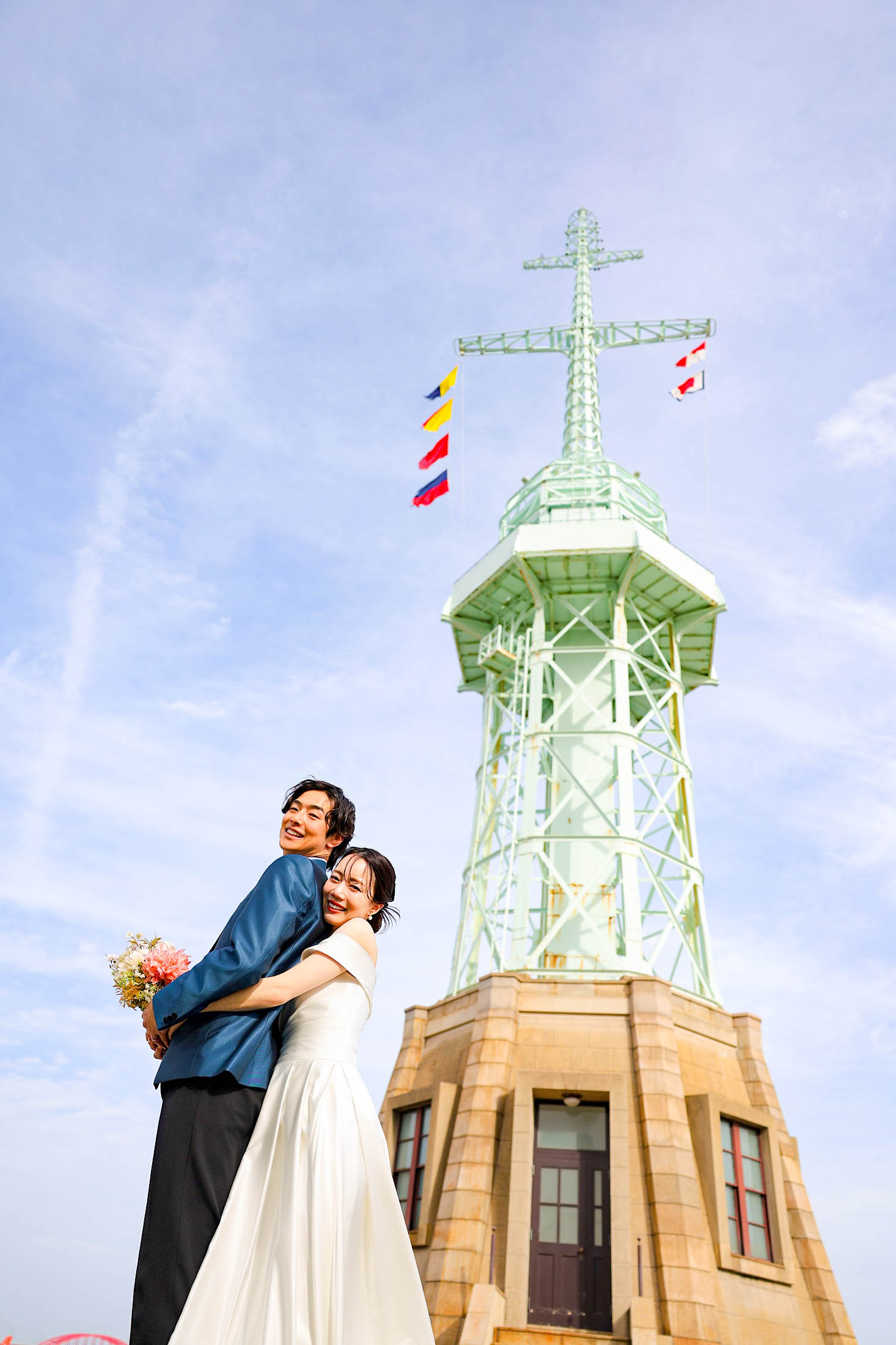 神戸ハーバーランドの旧神戸港信号所（旧新港第5突堤信号所）前でのフォトウエディング撮影。タキシードを着た新郎様にウエディングドレスを着た新婦様が後ろから笑顔で抱き着いている下アングルから撮影した写真。空と一緒に結婚の記念写真撮影