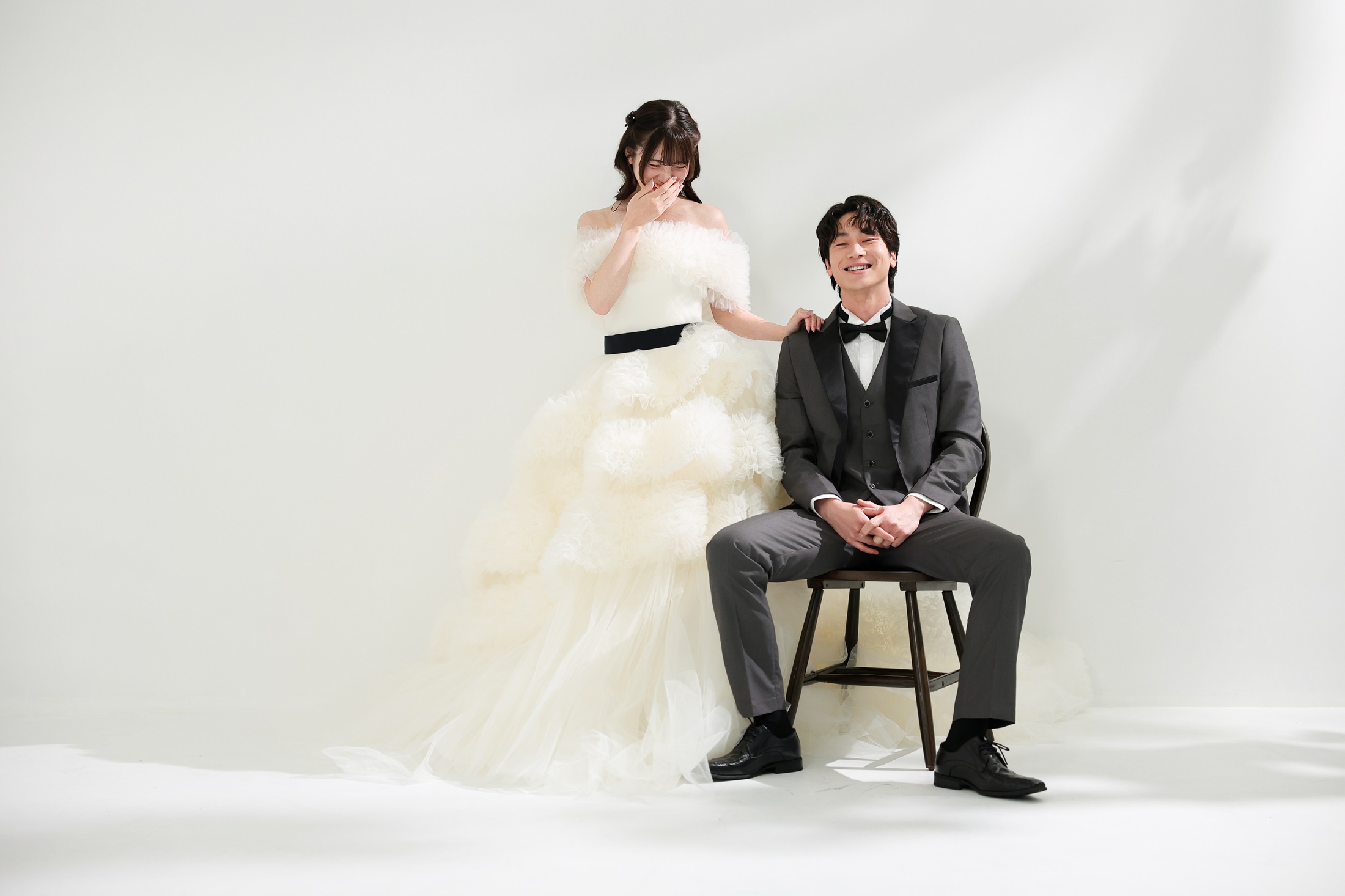 ウェディングドレスとタキシードでフォトウェディングの撮影をする新郎新婦様。柔らかな光が差し込む真っ白の壁を背景に、アンティーク調の椅子に腰を掛ける新郎様と横に立つ新婦様を撮影している写真。撮影店舗:ららテラスHARUMI FLAG店 住所:東京都中央区晴海五丁目2番31号 ららテラス HARUMI FLAG 3階
