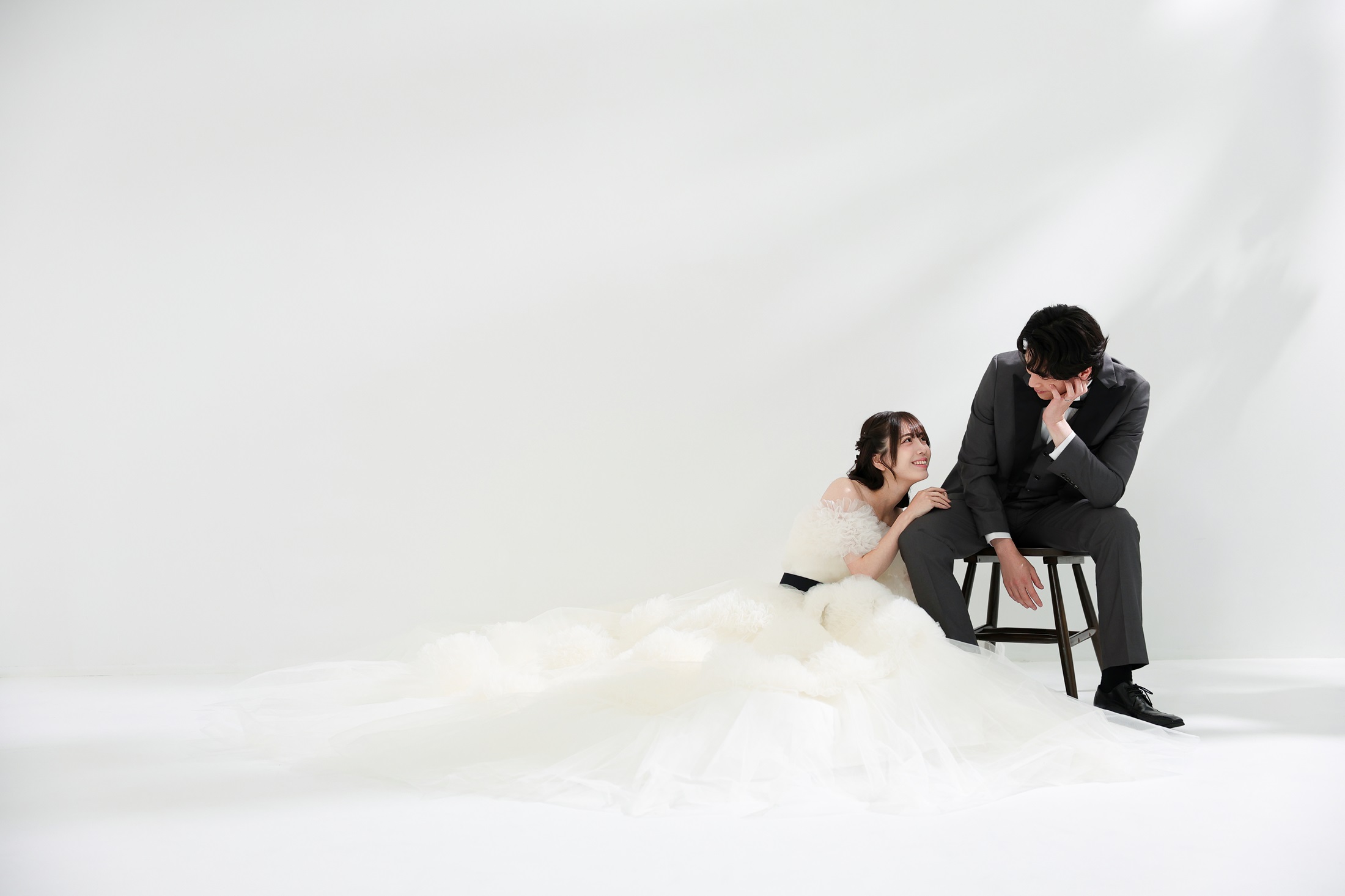 ウェディングドレスとタキシードでフォトウェディングの撮影をする新郎新婦様。真っ白の壁を背景に、椅子に腰掛ける新郎様とフロアにドレスを広げ腰を掛ける新婦様を撮影している写真。撮影店舗:ららテラスHARUMI FLAG店 住所:東京都中央区晴海五丁目2番31号 ららテラス HARUMI FLAG 3階