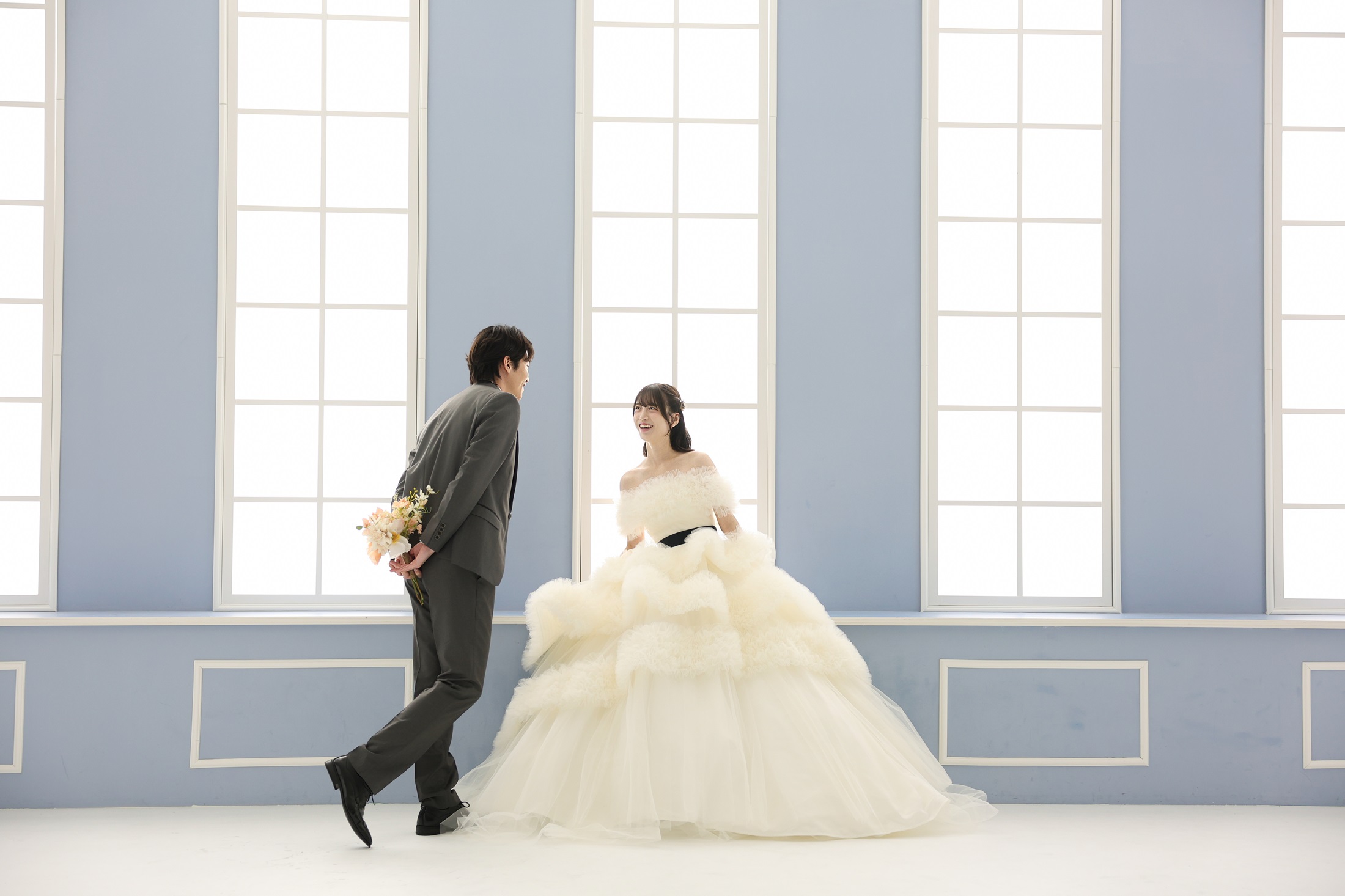 ウェディングドレスとタキシードでフォトウェディングの撮影をする新郎新婦様。柔らかなカーテンに水色の壁、大きな窓から注ぐ光がまるで宮殿のような背景で、新郎様が新婦様をエスコートするような笑顔の様子を撮影している写真。 撮影店舗:ららテラスHARUMI FLAG店 住所:東京都中央区晴海五丁目2番31号 ららテラス HARUMI FLAG 3階