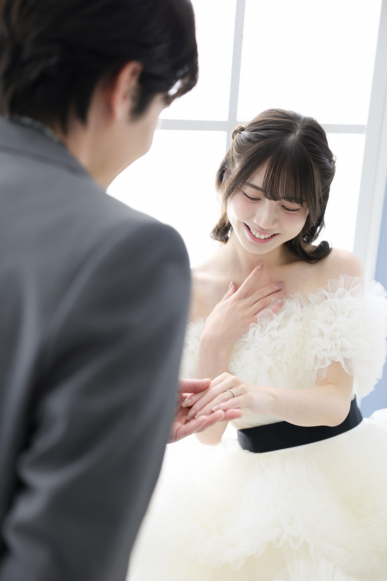 ウェディングドレスとタキシードでフォトウェディングの撮影をする新郎新婦様。柔らかな光が注ぐ窓を背景に、新郎様が新婦様の手を取りエスコートするような笑顔の様子を撮影している写真。 撮影店舗:ららテラスHARUMI FLAG店 住所:東京都中央区晴海五丁目2番31号 ららテラス HARUMI FLAG 3階