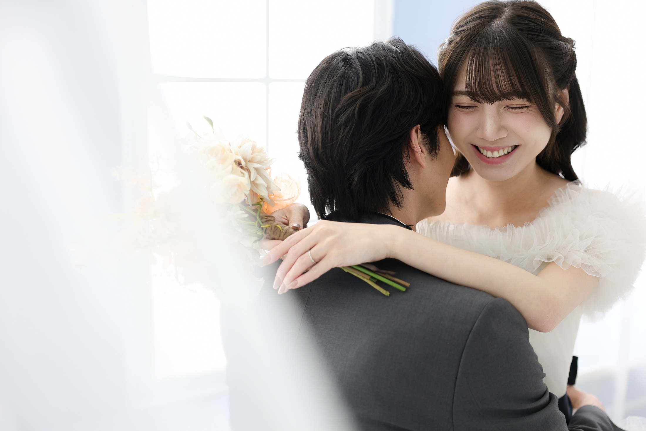 ウェディングドレスとタキシードでフォトウェディングの撮影をする新郎新婦様。柔らかな光に包まれる中、新婦様が新郎様に抱きつき笑顔の様子を撮影している写真。 撮影店舗:ららテラスHARUMI FLAG店 住所:東京都中央区晴海五丁目2番31号 ららテラス HARUMI FLAG 3階