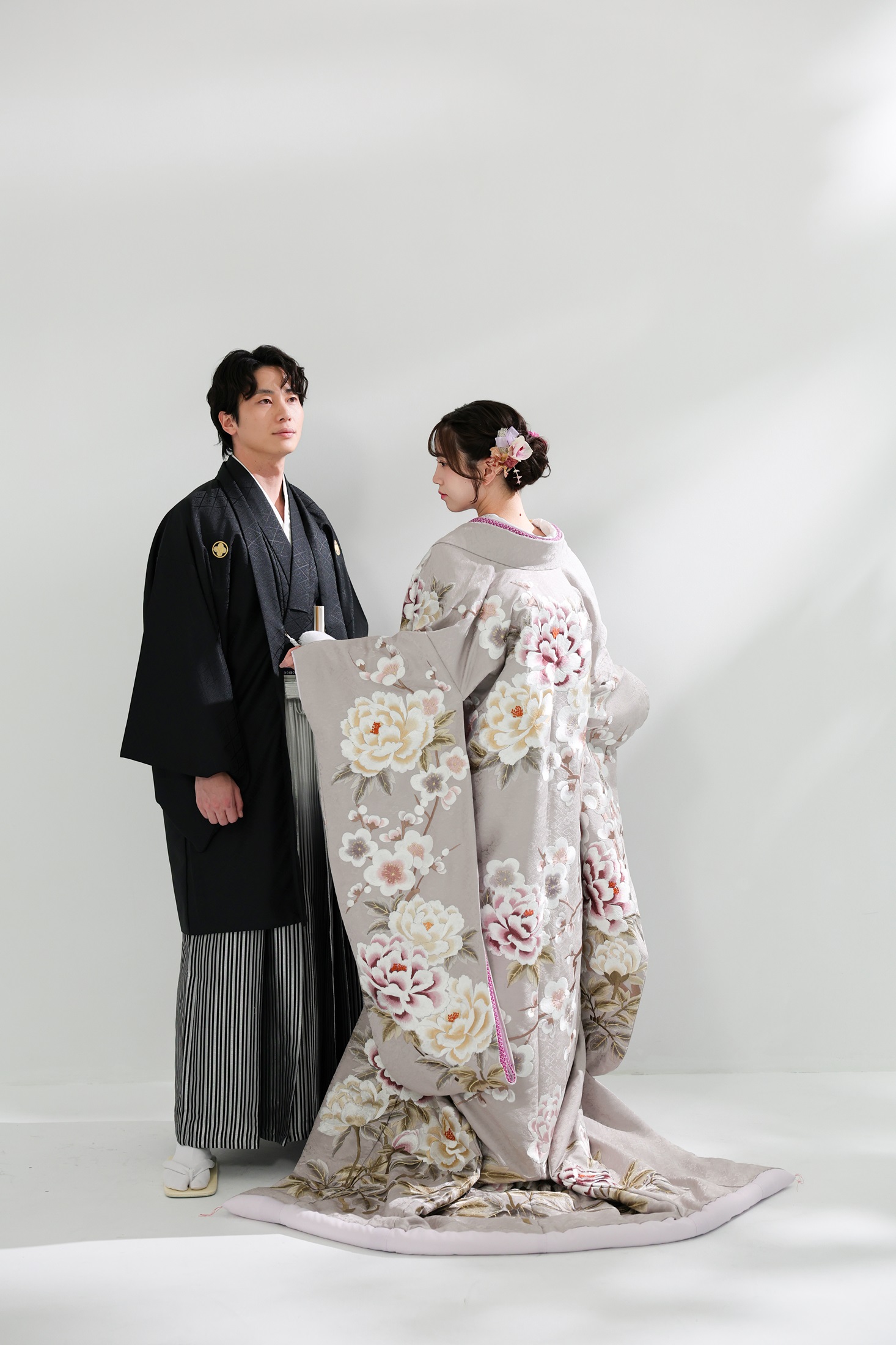 色打掛と袴でフォトウェディングの撮影をする新郎新婦様。柔らかな光が注ぐ真白の壁を背景に、後ろ姿の淡い色合いの色打掛が映える新婦様と、新郎様を撮影している写真。撮影店舗:ららテラスHARUMI FLAG店 住所:東京都中央区晴海五丁目2番31号 ららテラス HARUMI FLAG 3階