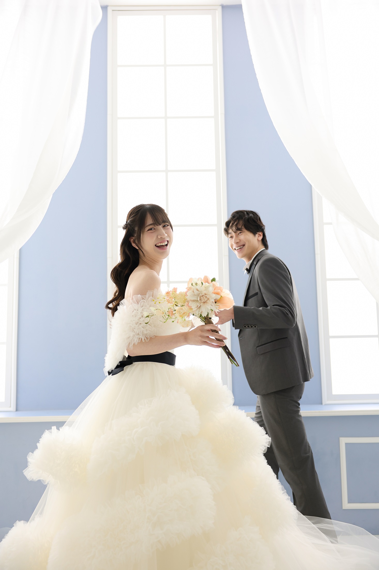 ウェディングドレスとタキシードでフォトウェディングの撮影をする新郎新婦様。柔らかなカーテンに水色の壁、大きな窓から注ぐ光がまるで宮殿のような背景で、新郎様が新婦様をエスコートするような笑顔の様子を撮影している写真。 撮影店舗:ららテラスHARUMI FLAG店 住所:東京都中央区晴海五丁目2番31号 ららテラス HARUMI FLAG 3階