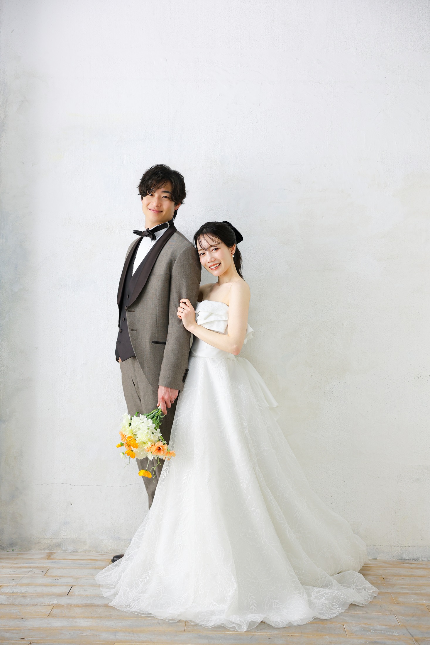 ウェディングドレスとタキシードでフォトウェディングの撮影をする新郎新婦様。真っ白な壁を背景に、ウェディングドレスが映える新婦様とオレンジ色のブーケを持つ新郎様の全身を撮影した写真。撮影店舗：神戸ハーバーランド店 住所：兵庫県神戸市中央区東川崎町1-5-5 ハーバーランド煉瓦倉庫