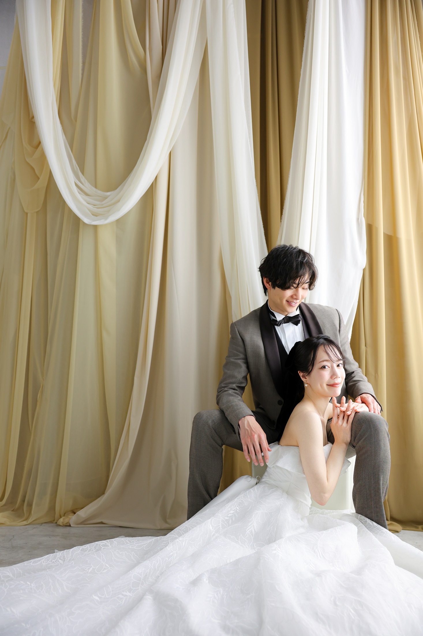 ウェディングドレスとタキシードでフォトウェディングの撮影をする新郎新婦様。柔らかでナチュラルなカーテンを背景に、新婦様がフロアに腰を下ろし、新郎に寄り添い、ドレスの裾が美しく広がる様子を撮影した写真。撮影店舗：神戸ハーバーランド店 住所：兵庫県神戸市中央区東川崎町1-5-5 ハーバーランド煉瓦倉庫