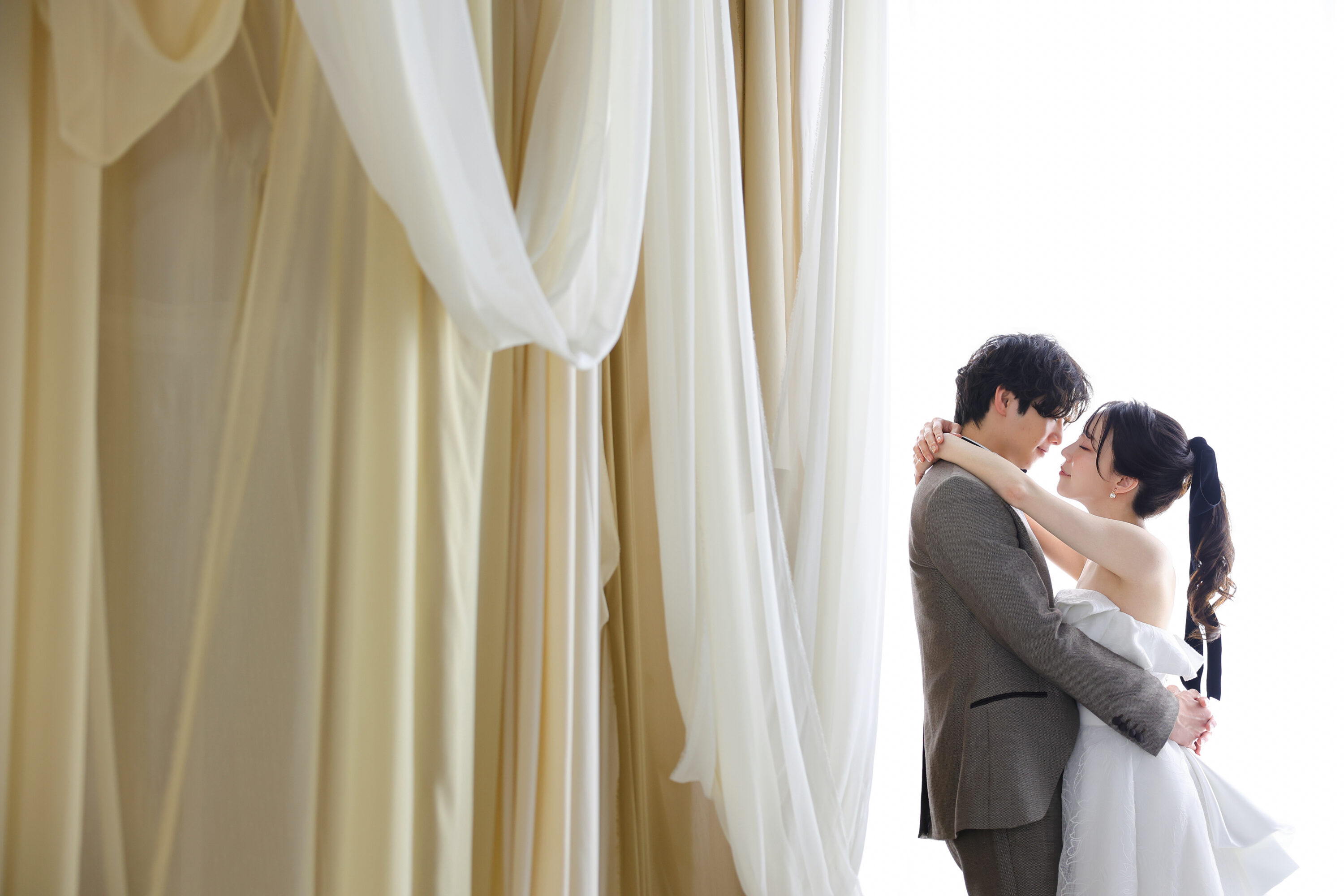 ウェディングドレスとタキシードでフォトウェディングの撮影をする新郎新婦様。柔らかでナチュラルなカーテンを背景に撮影した写真。撮影店舗：神戸ハーバーランド店 住所：兵庫県神戸市中央区東川崎町1-5-5 ハーバーランド煉瓦倉庫