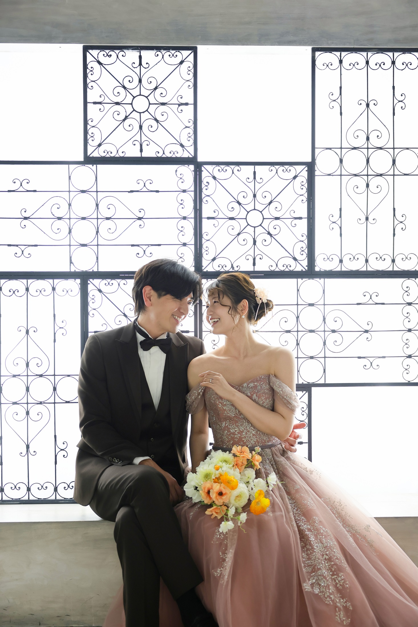 カラードレスとタキシードでフォトウェディングの撮影をする新郎新婦様。レトロな面格子のデザインを施した窓に腰かけ微笑み合う新郎新婦様を撮影した写真。撮影店舗：神戸ハーバーランド店 住所：兵庫県神戸市中央区東川崎町1-5-5 ハーバーランド煉瓦倉庫