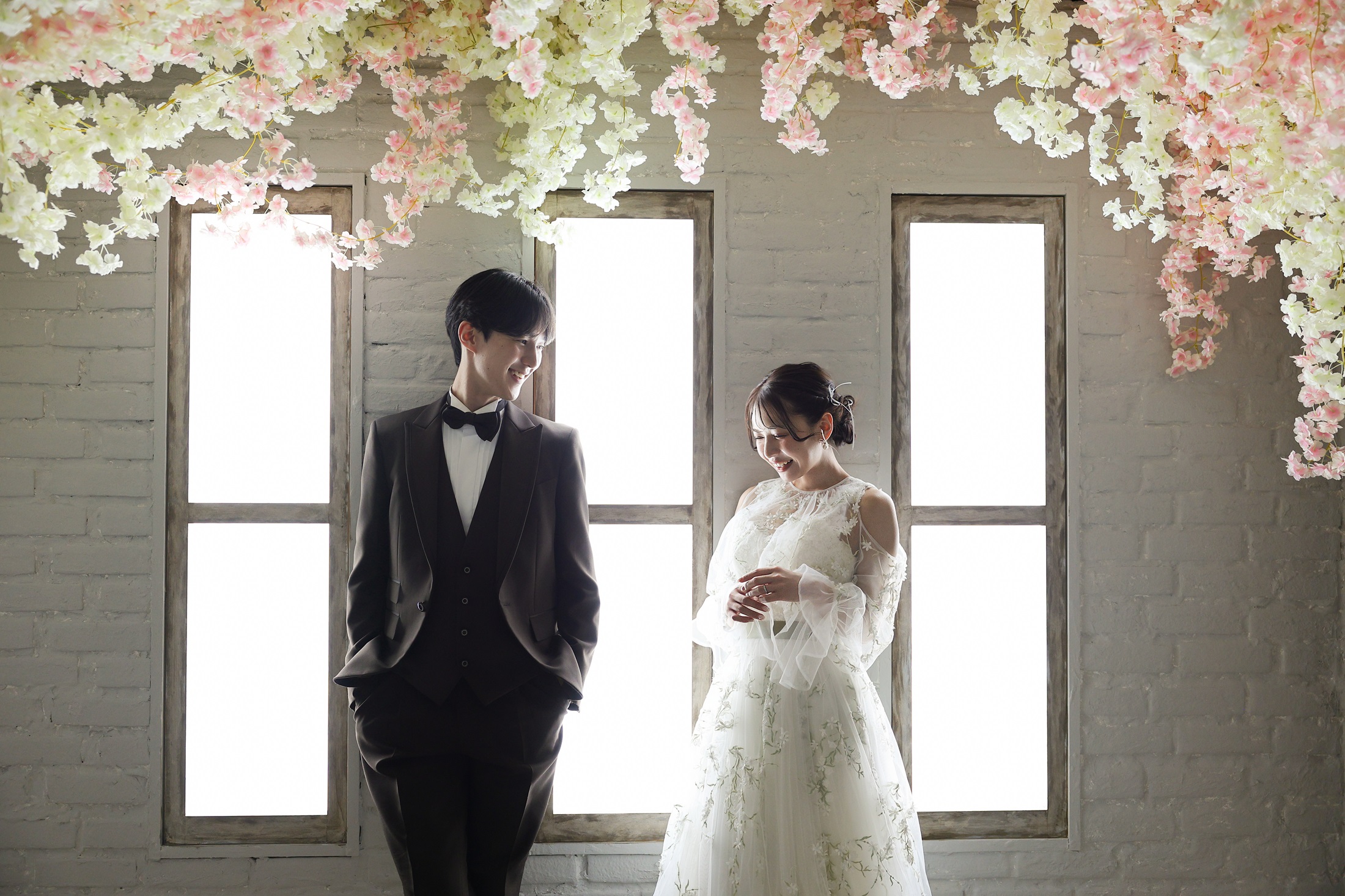 ウェディングドレスとタキシードでフォトウェディングの撮影をする新郎新婦様。天井から咲き誇るピンクと白の花々と、窓からの優しい逆光に包まれる中、二人並んで何気ない会話をしているようなナチュラルな写真。撮影店舗：神戸ハーバーランド店 住所：兵庫県神戸市中央区東川崎町1-5-5 ハーバーランド煉瓦倉庫