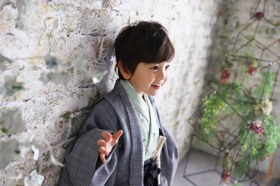 グレンチェックの羽織に淡いグリーンの着物を合わせた七五三の男の子が、白レンガの壁のそばで笑顔を見せる写真