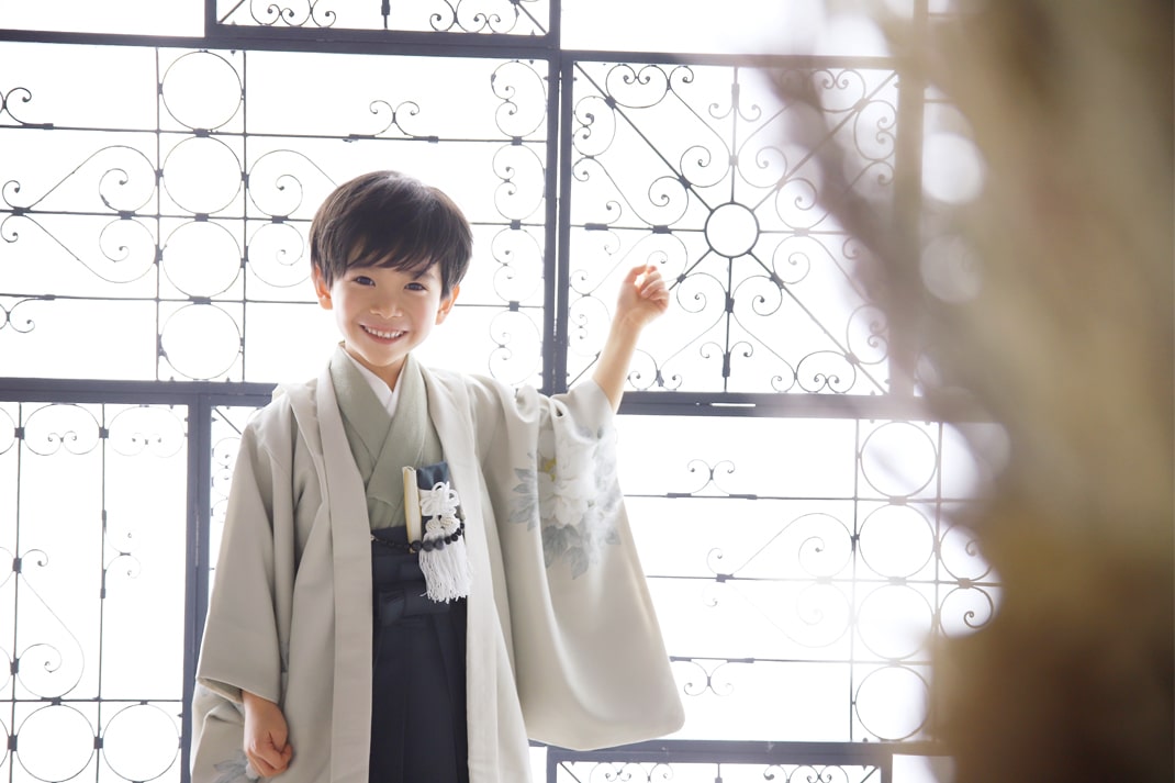 アイボリーの羽織と黒の袴を着用し、装飾的な窓を背景に笑顔を見せる男の子