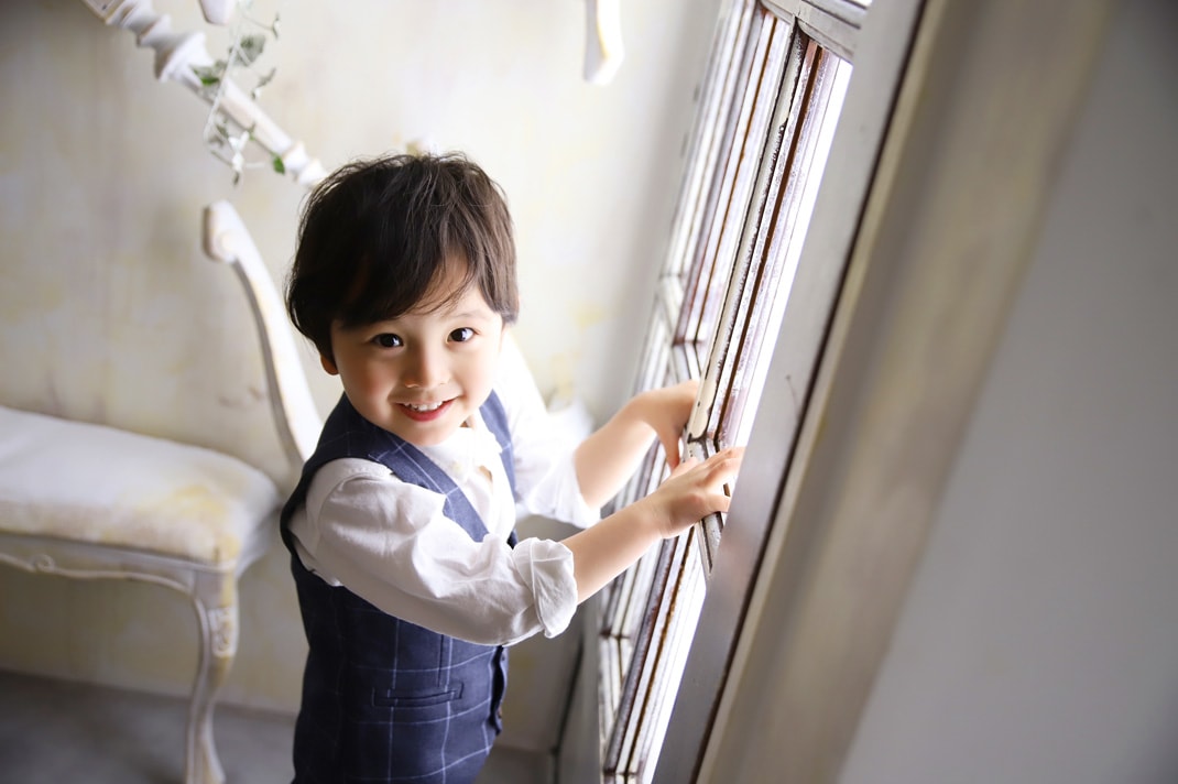 七五三の記念撮影で、白シャツにネイビーのチェック柄ベストを合わせた男の子が、大きな窓枠に手をかけて笑顔でこちらを見ている写真