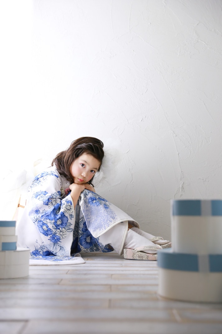 青い花柄の白い着物を着て床に座り頬に手を添える七五三の女の子の写真