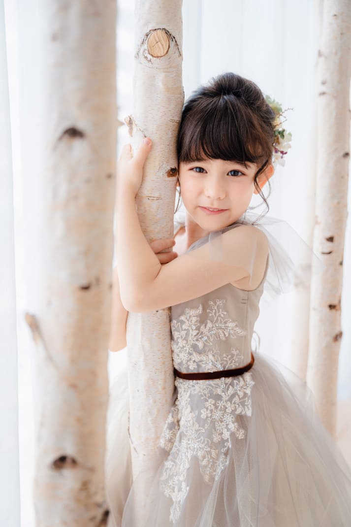 七五三の記念撮影で、グレー系のレースドレスを着た女の子が、白樺の木に寄り添いながら微笑んでいる写真