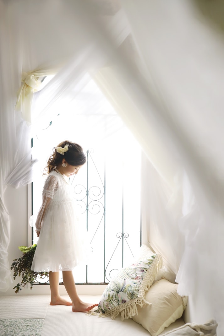 白いドレスを着て窓辺に立ち、足元を見つめる七五三の女の子の写真