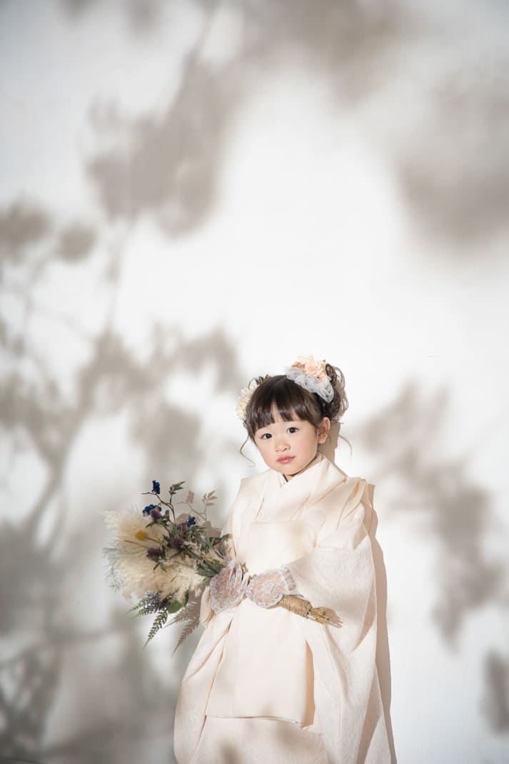 白い着物を着てブーケを持ち、木漏れ日のような影が映る壁の前に立つ女の子の写真