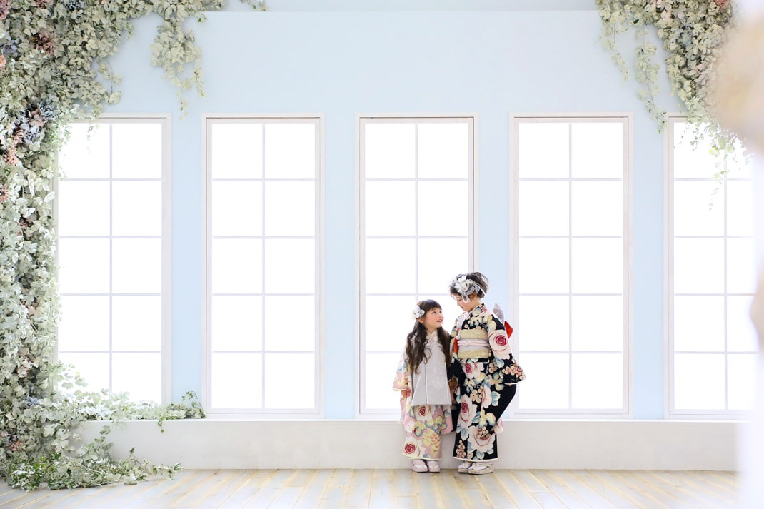 華やかな花柄の着物を着て、緑に囲まれた大きな窓の前で向き合う姉妹の写真