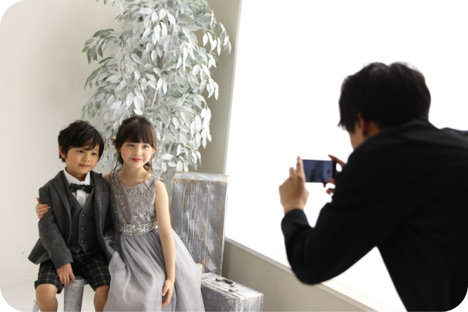 フォーマルな衣装の兄妹をスマホで撮影する保護者。スタジオでの自由撮影の様子