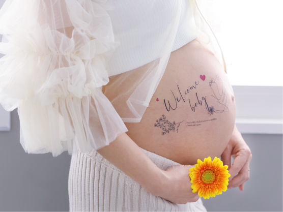 「Welcome baby」の文字や足形のマタニティシールを貼った大きなお腹のアップ。手には鮮やかなオレンジ色の花を持ったSTUDIO ARCのマタニティフォト