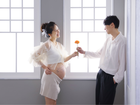 STUDIO ARCのマタニティフォト。窓際でパートナーから一輪の花を贈られ、マタニティシールを貼ったお腹とともに幸せそうに見つめ合う妊婦さん