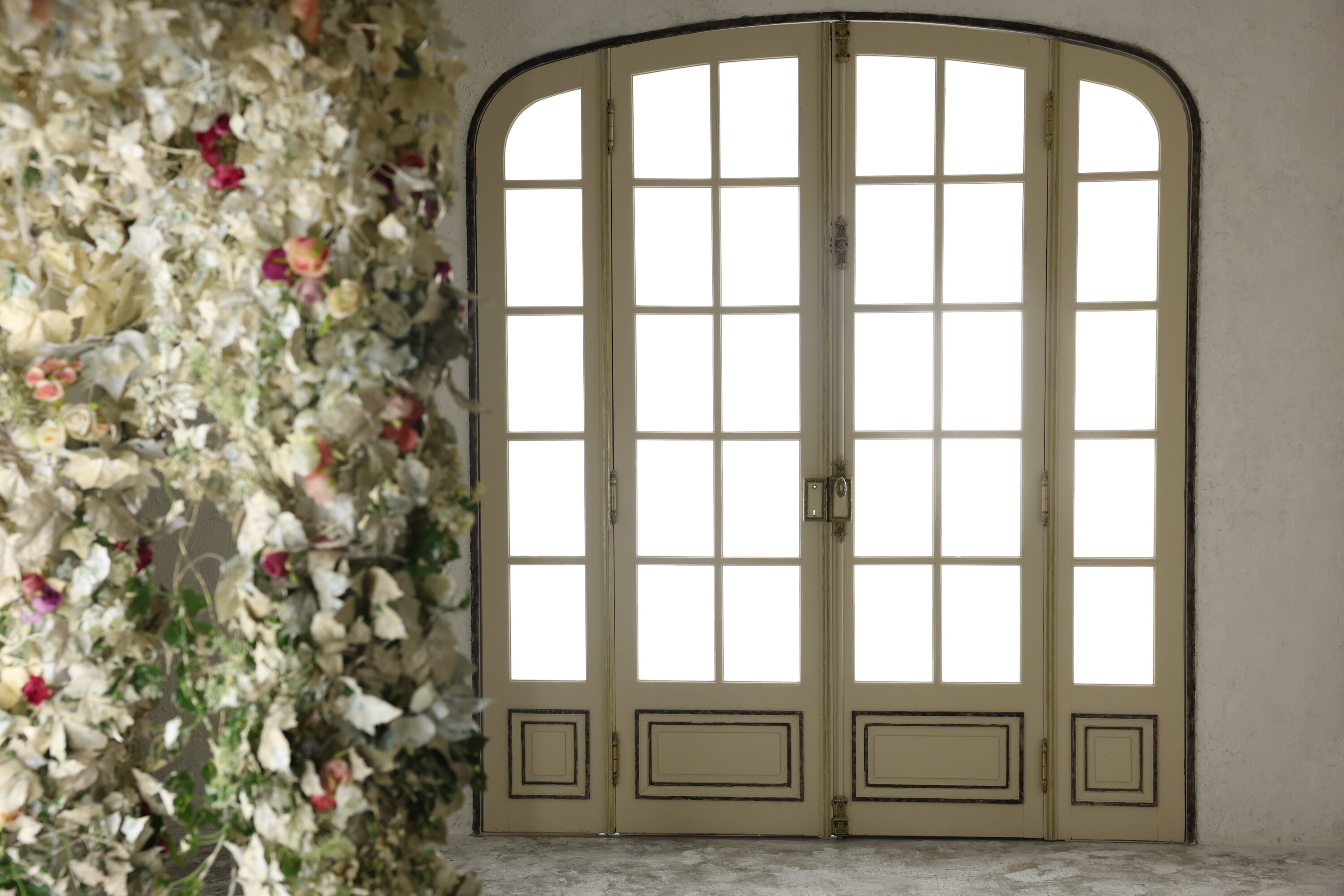 STUDIO ARC枚方店の撮影セット。グレージュの塗り壁に、上部がアーチ型になったアイボリーの格子窓付き両開きドアがあり、窓から白い光が差し込んでいる。ドアの下部には装飾パネル、中央にはアンティーク調の金色の金具がついている。画面左手前には、白い葉やピンク・赤・クリーム色のバラを重ねたボリュームのある花飾りが配置された、ヨーロッパ風のクラシカルな空間。