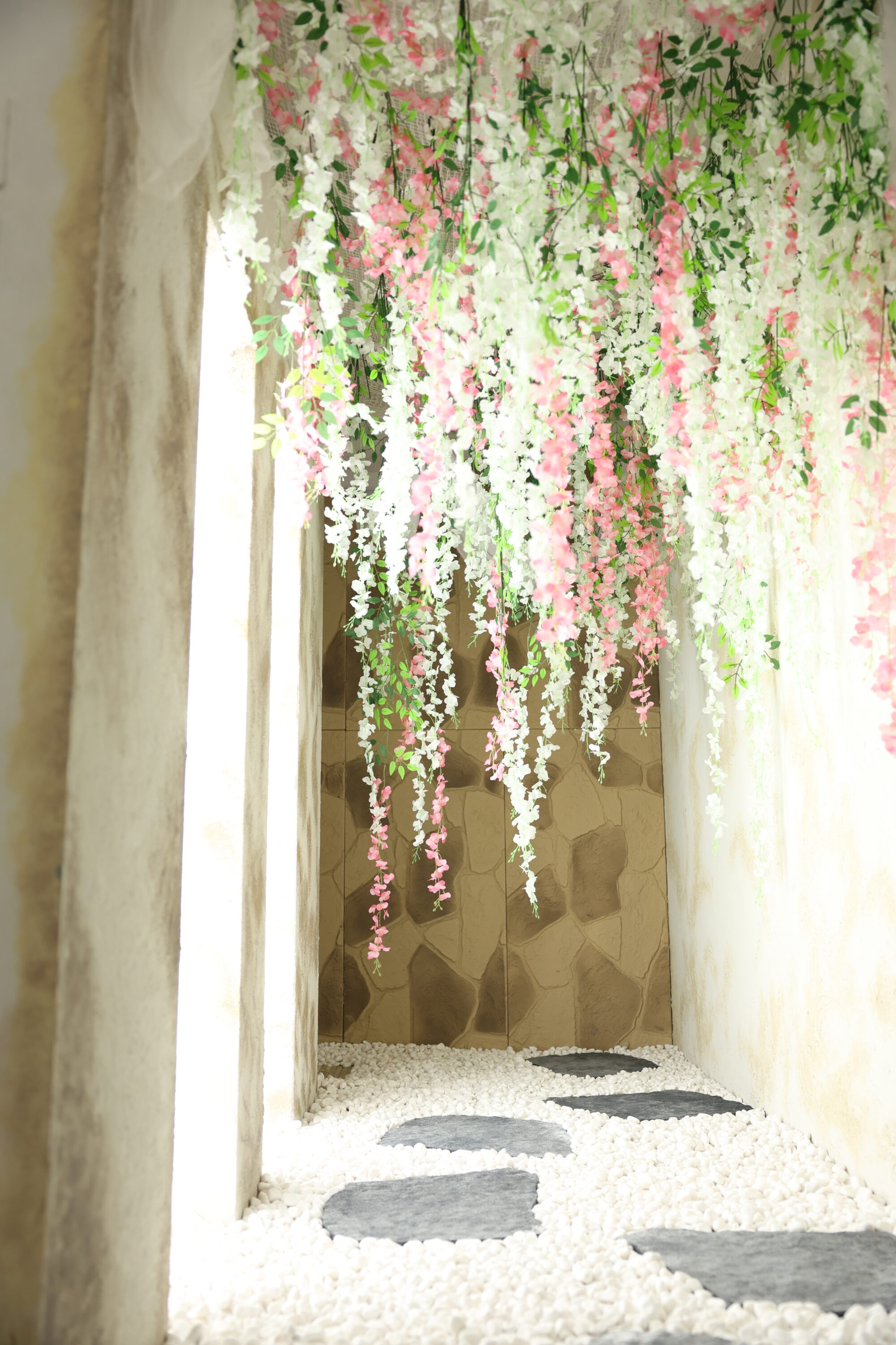 STUDIO ARC枚方店の撮影セット。天井一面から白とピンクの藤のような花飾りと緑の葉が垂れ下がり、足元には白い玉砂利の上にグレーの飛び石が奥へと続いている。突き当たりは石積み風の壁、両側はアンティーク調の白い壁で、花のトンネルをくぐるような和の雰囲気の空間。
