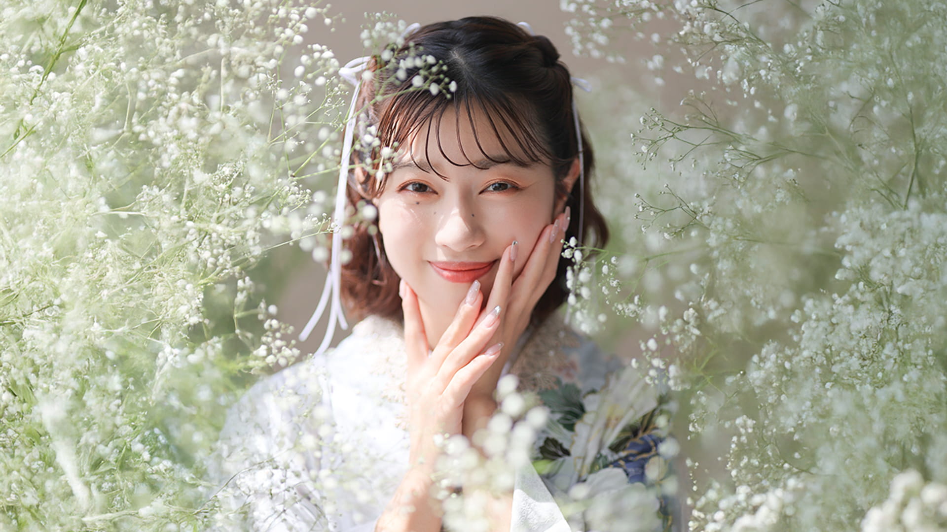 白く可憐なかすみ草に囲まれたスタジオで、ヘアリボンと上品な振袖を身につけた女性が手を頬に当てて微笑む成人式前撮りのポートレート（振袖アーク）