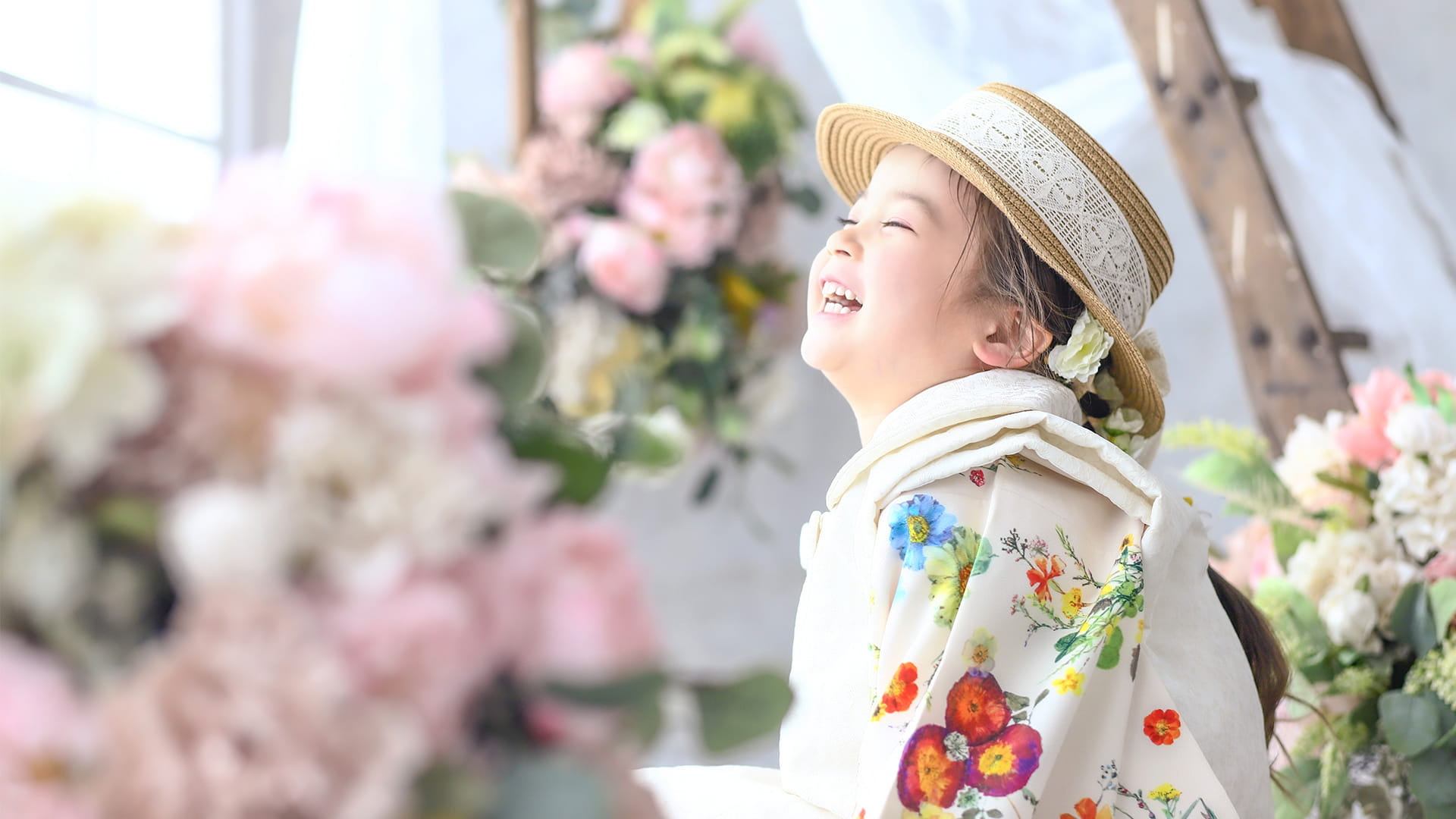 フォトスタジオで麦わら帽子と色鮮やかな花柄の和服を着て、たくさんの花飾りに囲まれながら笑顔を見せる女の子（七五三・キッズ撮影）
