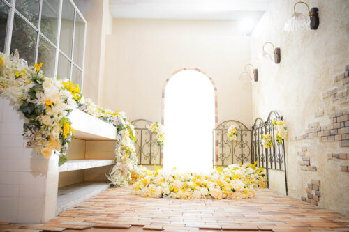 STUDIO ARCセンター北店のフォトブース。レンガタイルの床にアーチ型の入り口が続き、白×黄色の花々が通路一面と棚の装飾に飾られ、黒いシェードの壁付けランプとアイアンフェンスが上品なアクセントとなる、明るく華やかな撮影スペース。
