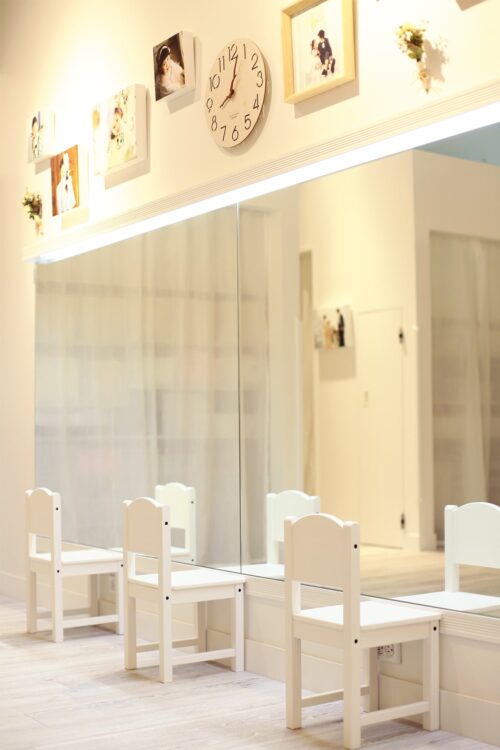 STUDIO ARCセブンパーク天美店のヘアメイクコーナー。大きな鏡の前に白い木製の子供用椅子が並び、上部の壁には作品写真や時計が飾られている、明るく清潔感のある店内スペース。