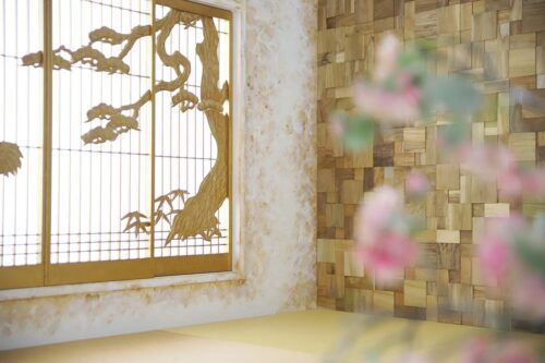STUDIO ARCセブンパーク天美店の和風の撮影セット。ゴールドの障子窓に松の木の意匠が描かれ、右側には木片を組み合わせたモザイク調の壁がある。手前には梅の花がぼかされて写り込んだ、格式高い和モダンなスタジオ空間。