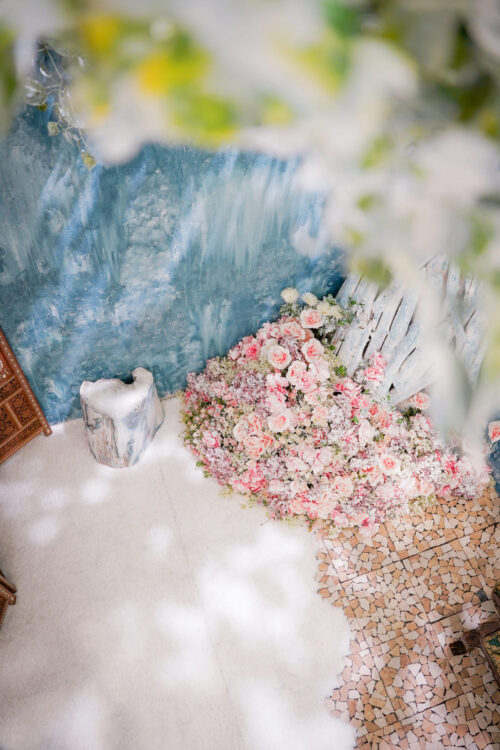 STUDIO ARC ららぽーと湘南平塚店の撮影セット。青いペイント調の壁の下に、ピンクや白の花がこんもりと敷き詰められ、白い切り株のスツールと、モザイクタイルの床が組み合わさった、絵画のようなおしゃれなスタジオ空間。