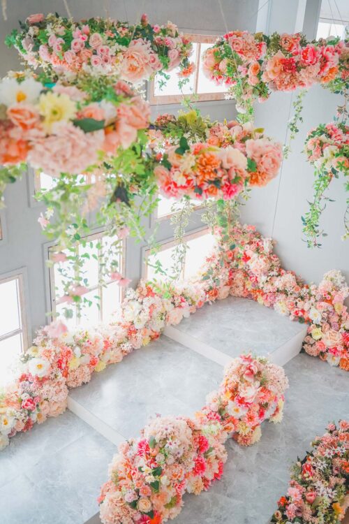 STUDIO ARC 錦糸町PARCO店の撮影セット。ピンクやオレンジ、白の花々が天井から吊るされ、グレーの大理石調のステージにも花が咲き誇る、華やかでフラワーガーデンのようなスタジオ空間。大きな窓から自然光が差し込んでいる。