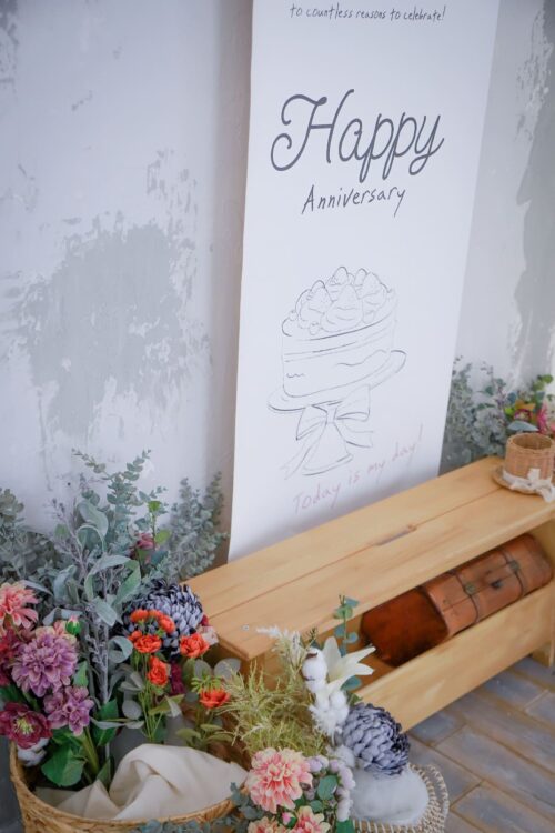 STUDIO ARCイオンモールくみやま店の撮影セット。「Happy Anniversary」の手書き風文字とケーキのイラストが描かれたポスターが壁に立てかけられ、木製のベンチとアンティーク調のトランク、色とりどりの花のバスケットが配された、温かみのあるナチュラルなスタジオ空間。