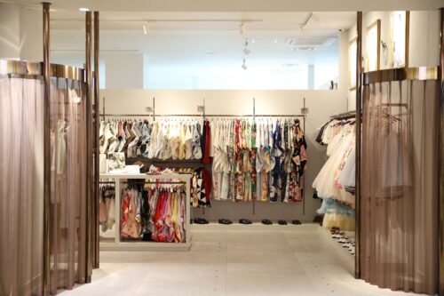 STUDIO ARCららぽーと門真店の店内。七五三の着物やドレスがカラフルに並ぶ衣装レンタルコーナーで、豊富な柄の着物、白いフォーマルドレス、靴も種類豊富に揃っている。両サイドには落ち着いたブラウン系のカーテンの試着スペースがある、明るく清潔感のある店内。