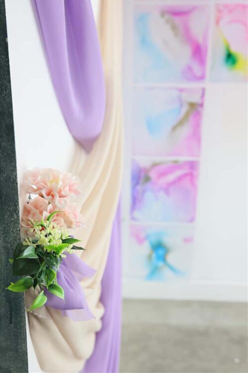 STUDIO ARCららぽーと甲子園店の入り口に飾られた紫とベージュのドレープ装飾。淡いピンクの造花とグリーンを紫のリボンでまとめた花飾りが添えられ、背景には水彩画風のアートパネルが並ぶ、七五三撮影用の華やかな空間演出。