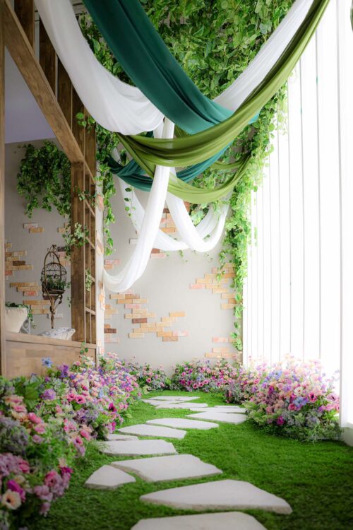 STUDIO ARCおおとり店の撮影セット。人工芝の小道に敷石が並び、両側にピンクや紫の花がふんだんに咲き誇る。天井からはグリーンの布と白、深緑、オリーブ色のドレープが垂れ下がる、庭園のようなナチュラルで華やかなスタジオ空間。