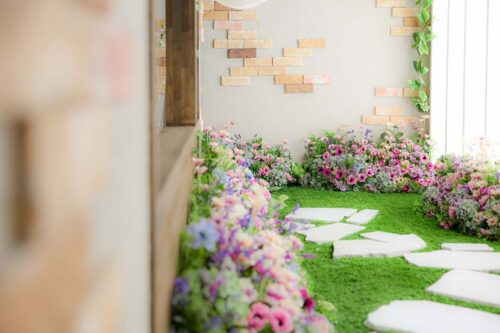 STUDIO ARCおおとり店の店内装飾。人工芝の床にピンクや紫、白の花々が縁取るように並び、白い飛び石が通路として配置されている。壁面にはレンガ調のタイルとグリーンの装飾があしらわれている
