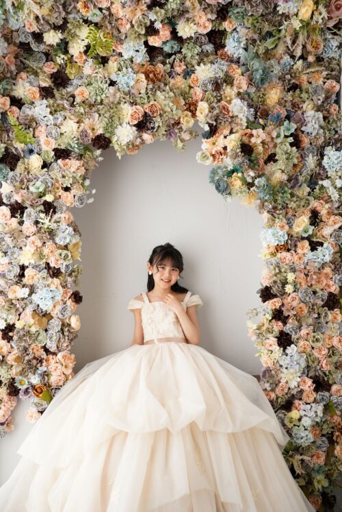 ららぽーと立川立飛店で撮影された、花のアーチの下に立つ女の子の記念写真。淡いベージュ色のオフショルダードレスを着て微笑んでおり、背景には色とりどりのバラやあじさいで作られた大きなフラワーアーチが飾られている
